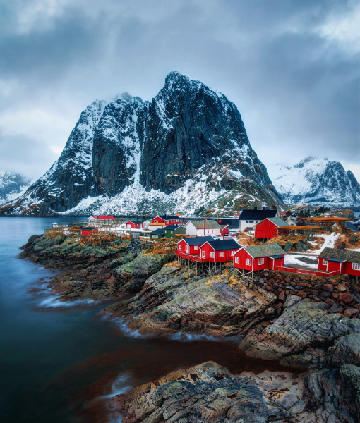 Snowy mountain and rocky coastline with colorful red houses clustered along the shore by calm water, small fishing village scene in winter - Aufgang Travel