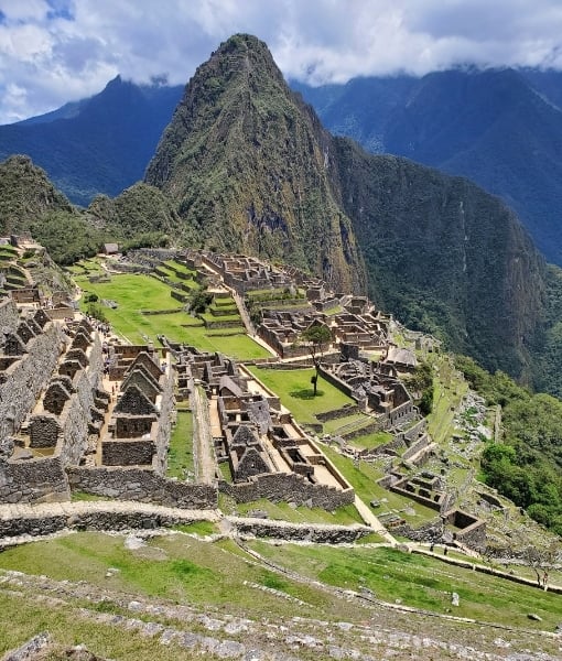 Ancient Inca citadel of Machu Picchu with stone terraces and stairways on a green hillside, Huayna Picchu looming in the background under a blue sky. - Aufgang Travel