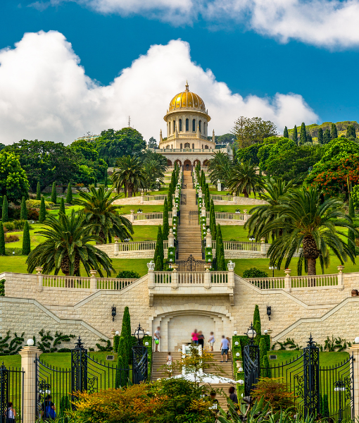 Wide stone staircase flanked by palm trees ascends a manicured garden toward a gold-domed building on a hill, with iron gates at the foreground. - Aufgang Travel