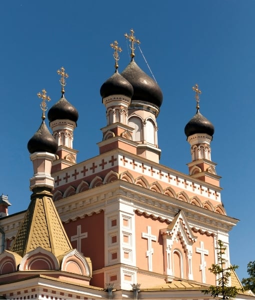 Ornate pink Orthodox church with white decorative detailing, black onion domes topped with gold crosses against a blue sky. - Aufgang Travel