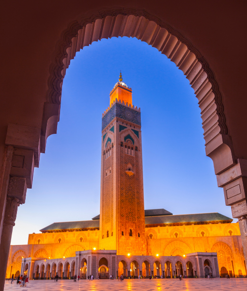Tall minaret framed by an ornate arch, with golden-lit mosque arches and a blue twilight sky in the background. - Aufgang Travel