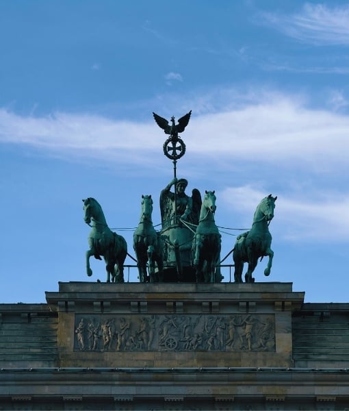 Quadriga statue atop a neoclassical gate: four horses pulling a chariot with a central figure, against a bright blue sky. - Aufgang Travel