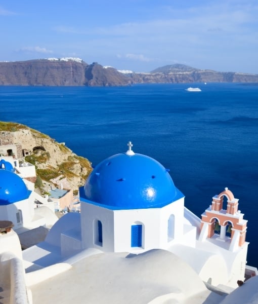 White-washed buildings and a vivid blue dome in Santorini, with the Aegean Sea and coastline in the background. - Aufgang Travel
