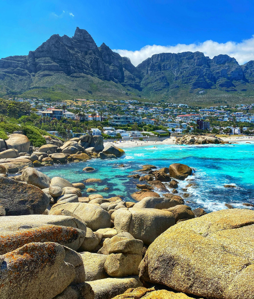Rocky beach with large granite boulders, bright turquoise water, and a town at the shoreline with towering mountains in the background. - Aufgang Travel