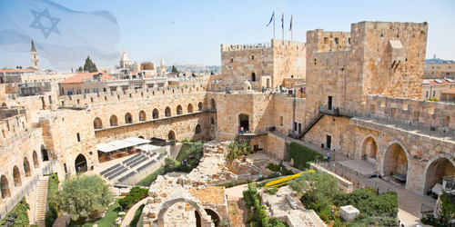 Panoramic view of a stone fortress with arched colonnades, towers, and a garden courtyard; an Israeli flag flies above. - Aufgang Travel