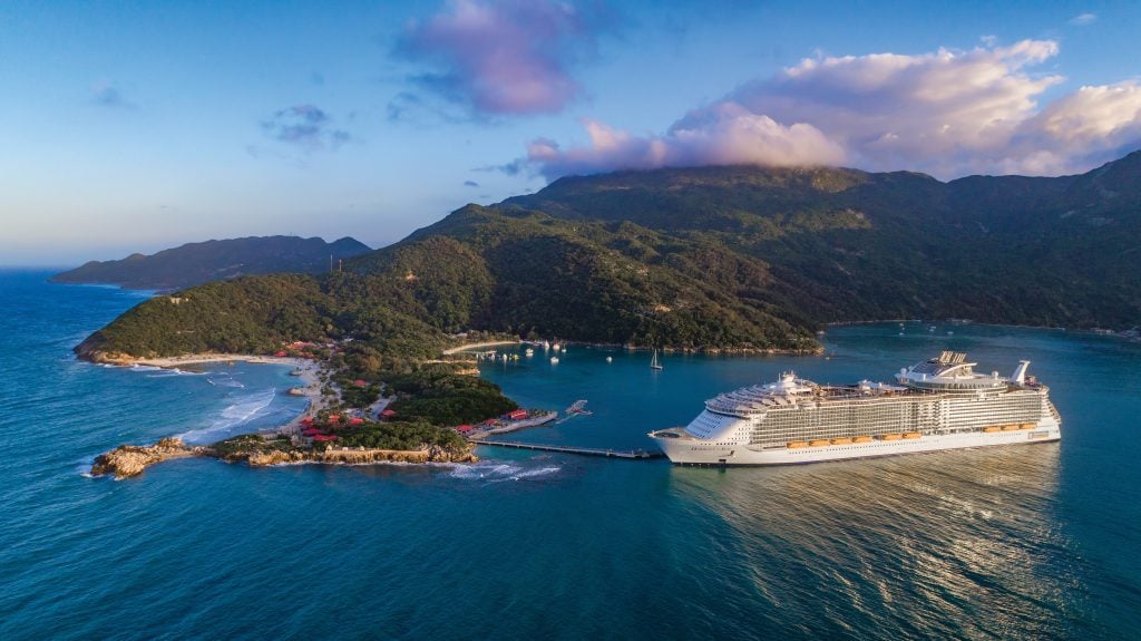 Large white cruise ship docked in a blue bay beside a forested tropical island and green mountains in the distance.