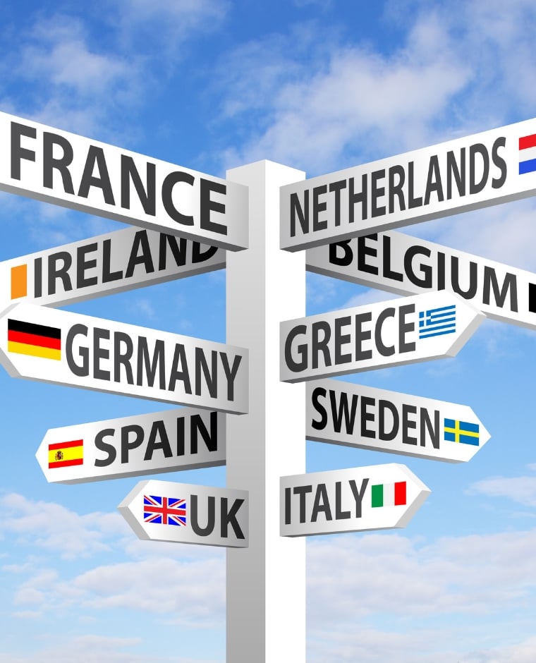 Signpost with arrows pointing to European countries and small flags (France, Ireland, Germany, Spain, UK, Netherlands, Belgium, Greece, Sweden, Italy) against a blue sky. - Aufgang Travel
