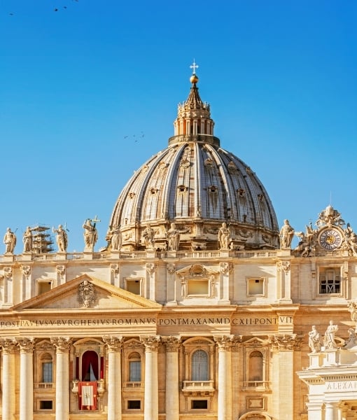 Front façade of St. Peter's Basilica with a large central dome and row of statues against a clear blue sky. - Aufgang Travel