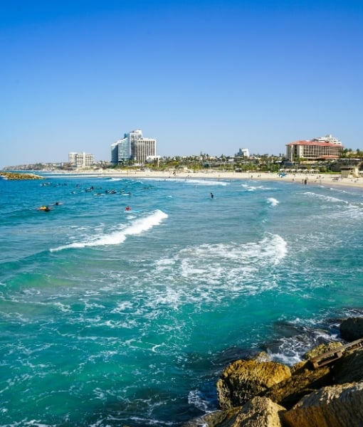 Sunny beach scene with turquoise water, waves, and people swimming near a rocky shore with a city skyline in the background on a clear day. - Aufgang Travel