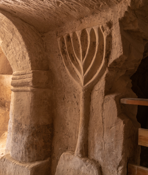Carved menorah on a rough stone wall inside a dim cave or tomb. - Aufgang Travel