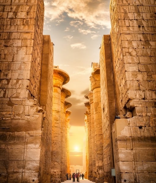 Ancient stone temple corridor with tall columns on both sides and a sunset glow at the end, with visitors walking through. - Aufgang Travel