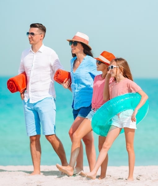 Family of four walks along a sunny beach with towels and a turquoise inflatable ring, sea in the background. - Aufgang Travel
