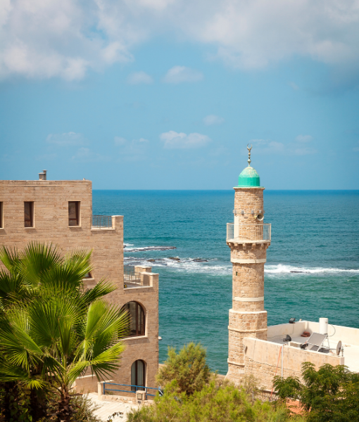 Coastal scene with beige stone buildings and a round minaret-style tower with a green dome by the blue sea. - Aufgang Travel