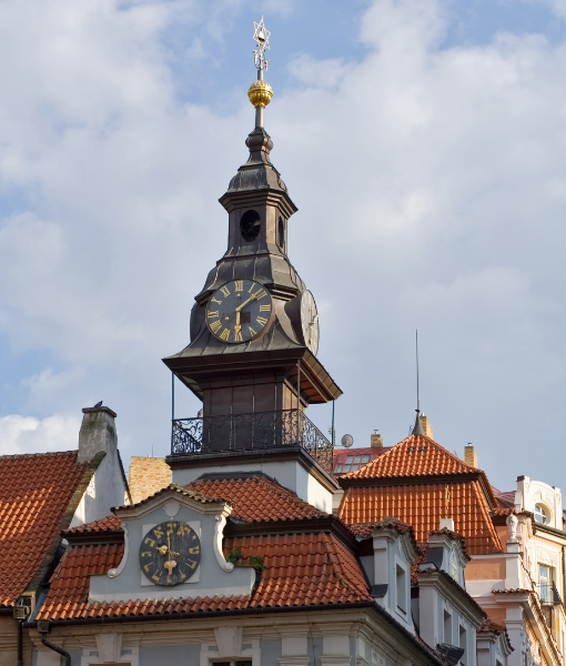 Historic clock tower rising above a red-tiled roof, with gold clock faces and a weather vane against a blue sky. - Aufgang Travel