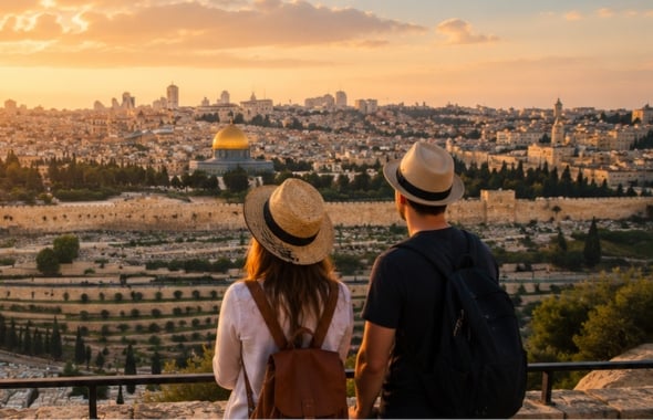 Two tourists with backpacks and hats stand at a viewpoint, gazing at Jerusalem’s skyline at sunset, with the Dome of the Rock visible in the distance.