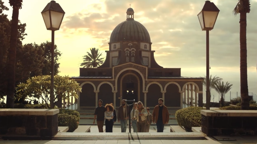 Group of six people walking toward a domed church at sunset, palm trees flanking a stone plaza by the sea in the background