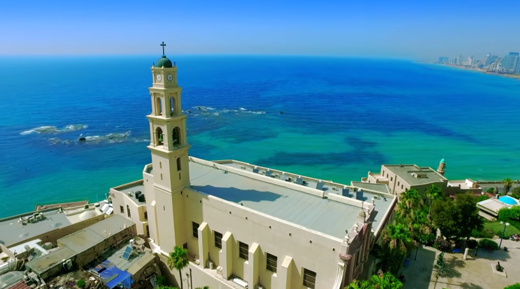 A tall tan church bell tower along a coastline with bright blue-green ocean and a distant city skyline on the right.