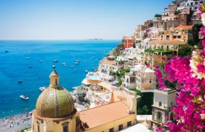 Colorful hillside town along blue sea with a tiled-domed church, boats in the harbor, and pink bougainvillea in the foreground.